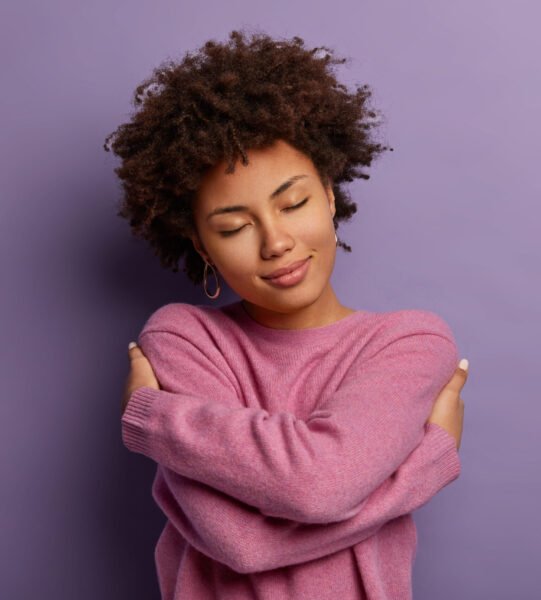 Romantic woman embraces herself, keeps arms crossed over body, touches shoulders, enjoys coziness, wears sweater, tilts head, has high self esteem, loves herself, isolated on purple background